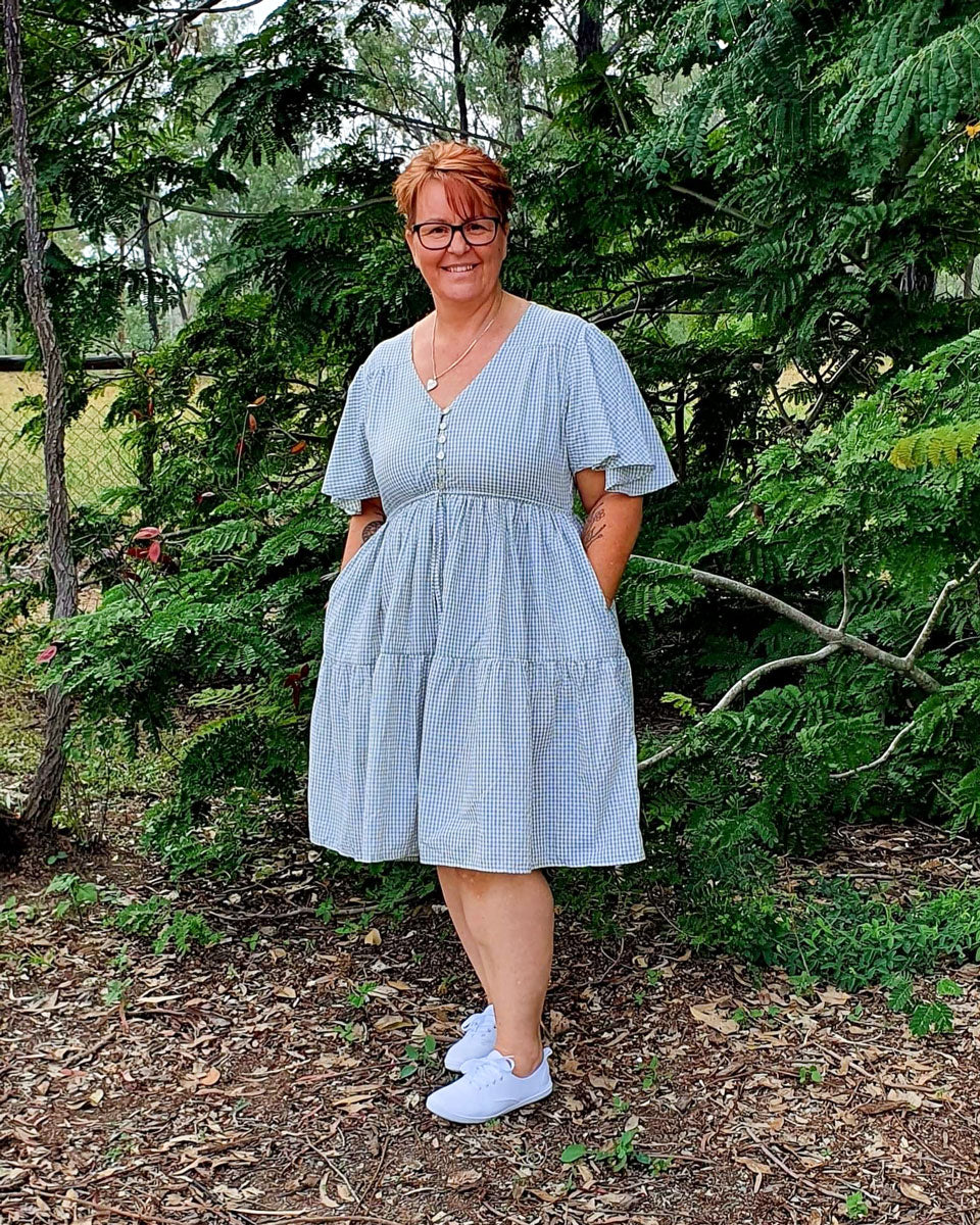 A woman with short hair and glasses wears a cute light-blue, knee-length v-neck dress. She smiles, her hands obscured deep in her pockets.