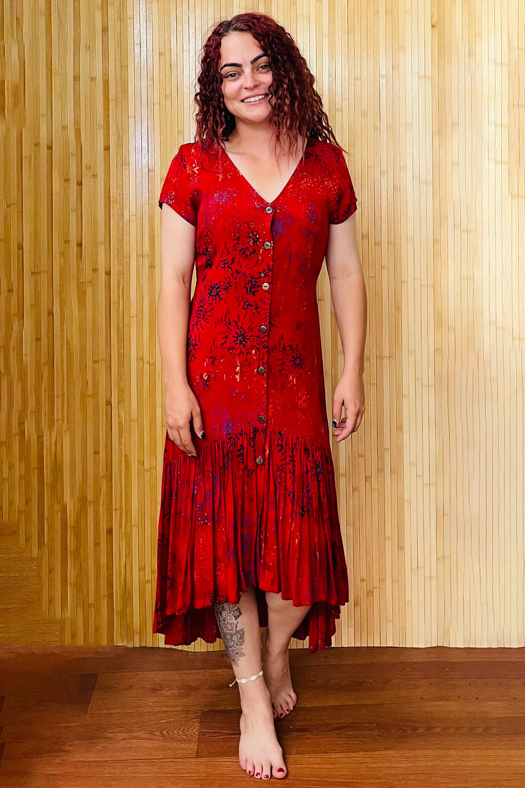 A smiling woman wears a v-neck red-patterned dress, with buttons down the front, short sleeves, and a gathered hi-low hem, which starts mid-calf at the front