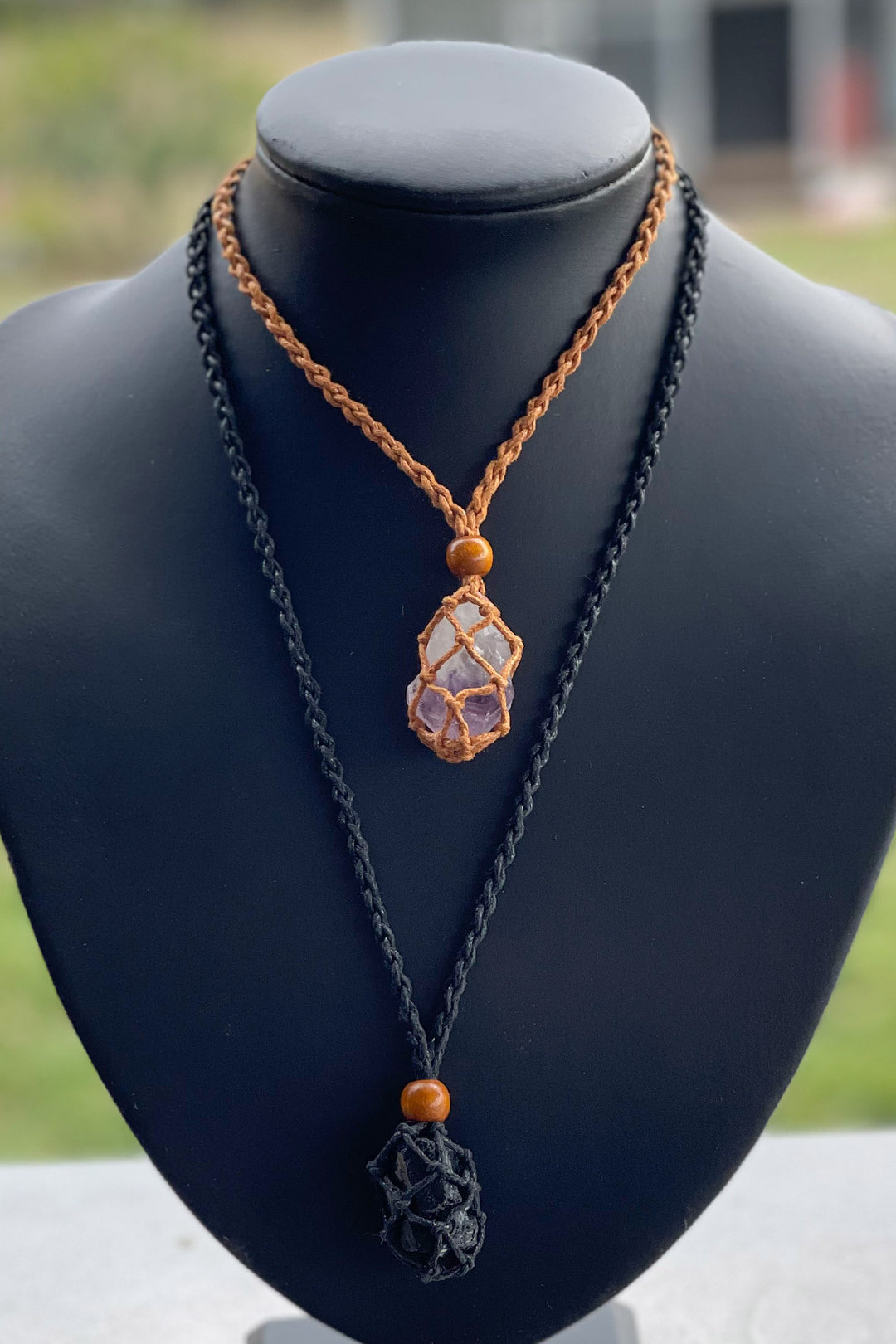 A short brown macrame necklace holding a piece of amethyst, worn with a long black version holding a black crystal. 